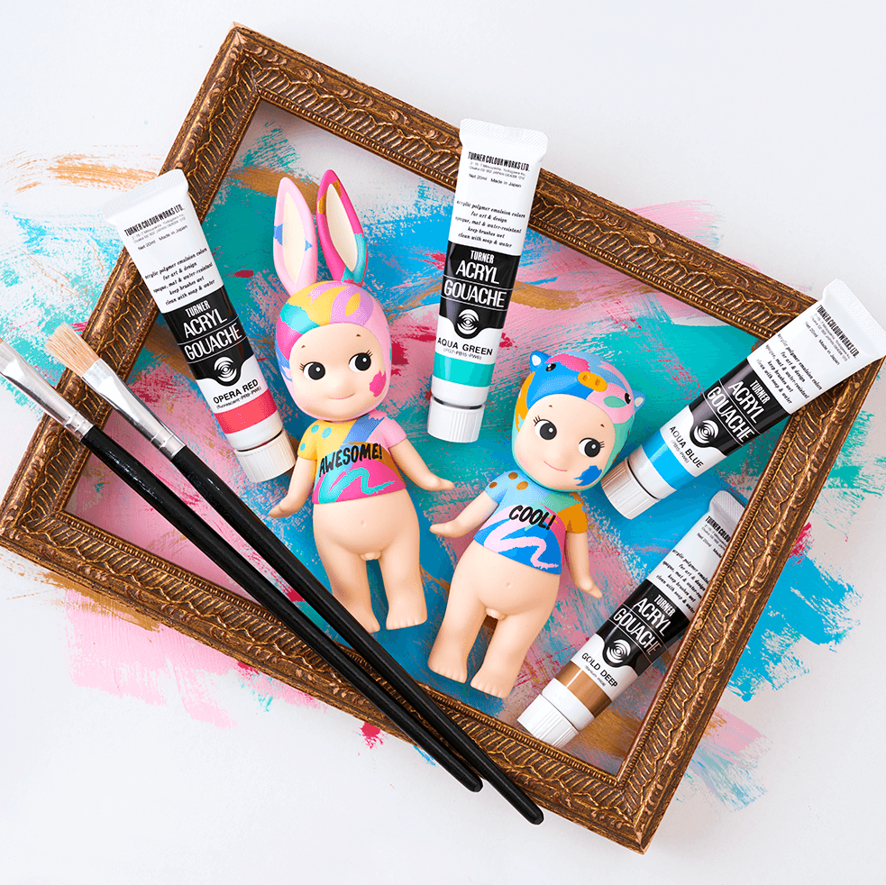 Two small dolls with colorful outfits inside a decorative frame with art supplies.