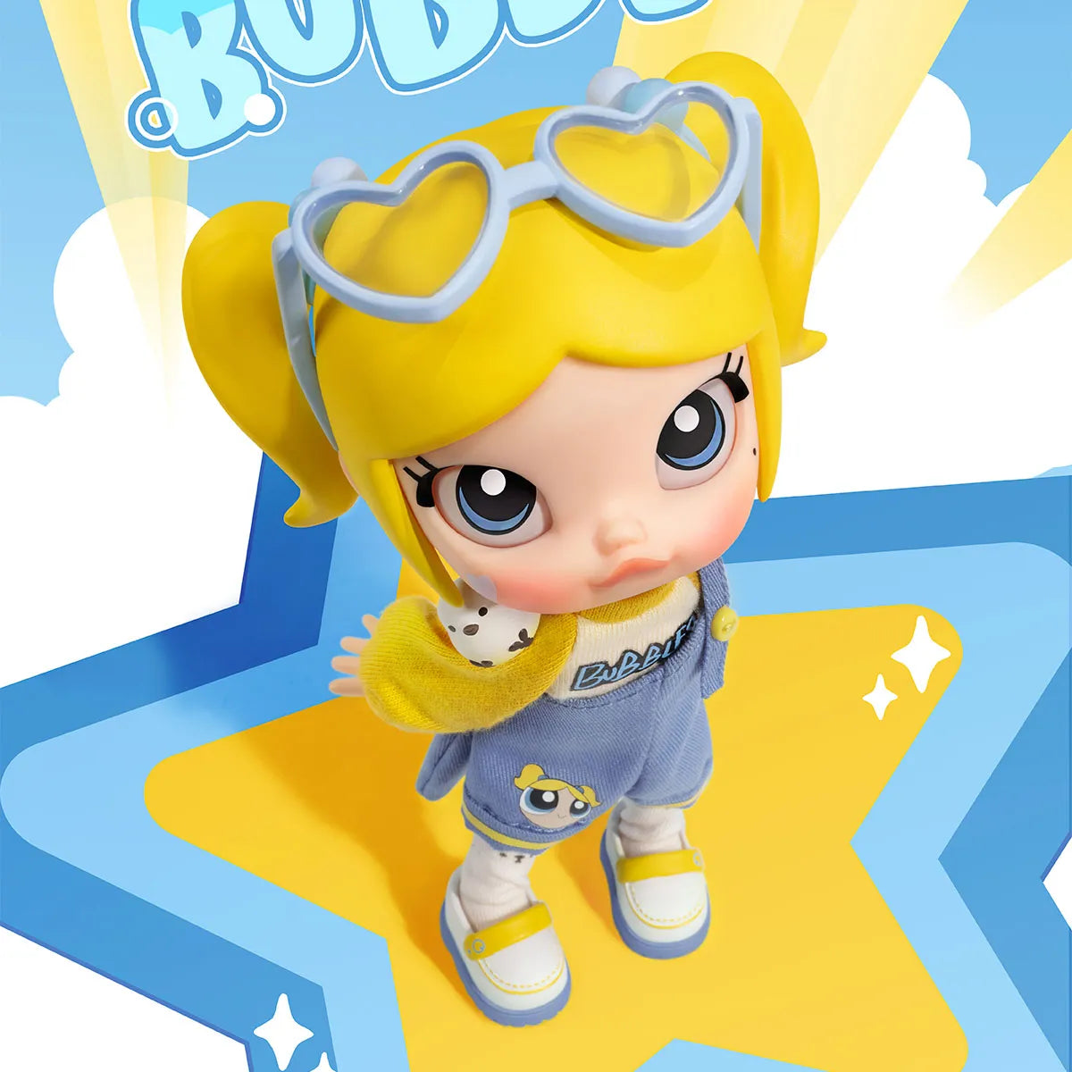 A stylized collectible doll or figurine with large blue eyes, yellow pigtails, heart-shaped goggles, a yellow top, and denim overalls, posed on a blue and yellow star-shaped graphic background.