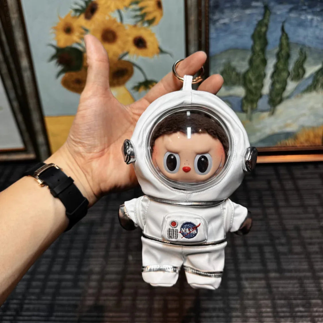 Keychain in the shape of a person in an astronaut suit held by a hand with Van Gogh's 'Sunflowers' and 'Cypress' in the background.