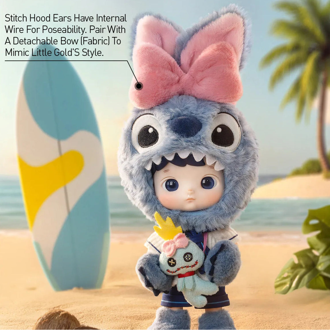 A plush doll dressed in a blue stitch costume with a pink bow on its head stands on a sandy beach. The doll holds a small white plush character with a yellow crown.