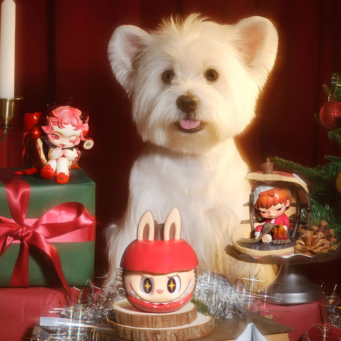 White dog with festive decorations including a red gift box, snow globe, and small figurines on a dark background.
