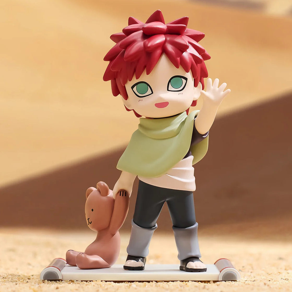 A chibi-style figurine of Gaara from Naruto standing in a desert scene, holding a small teddy bear. The figure has spiky red hair, green eyes, a light green scarf, a white shirt, dark gray pants, and black sandals, set against a blurred sandy background.