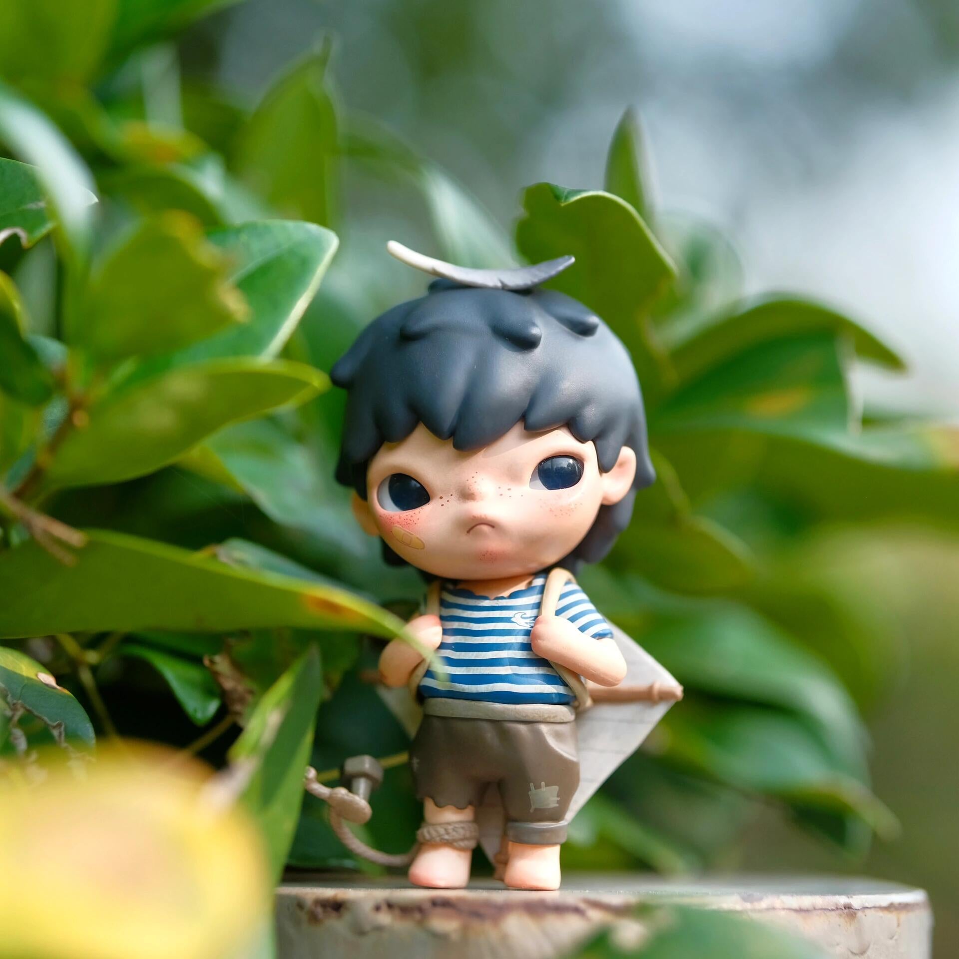 Hirono Kite Beijing Swift Figurine of a child with black hair and a striped shirt standing among green leaves.