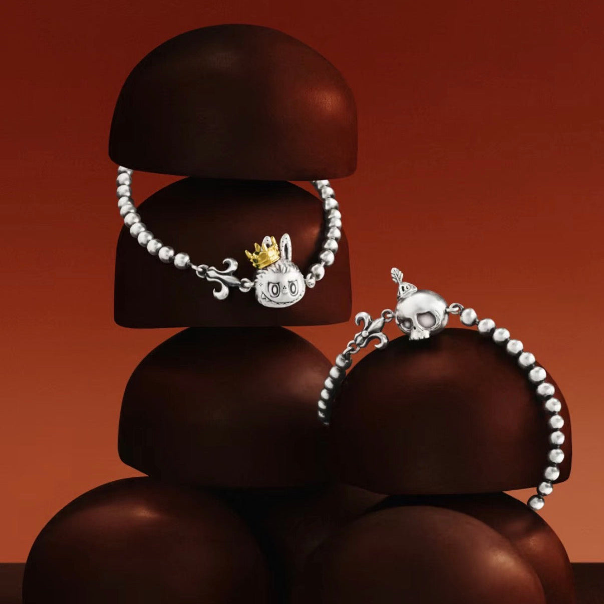 Two silver beaded bracelets displayed on dark brown sculptural stands against a warm orange-brown gradient background. One bracelet features a rabbit charm with a small gold crown, and the other features a skull charm.