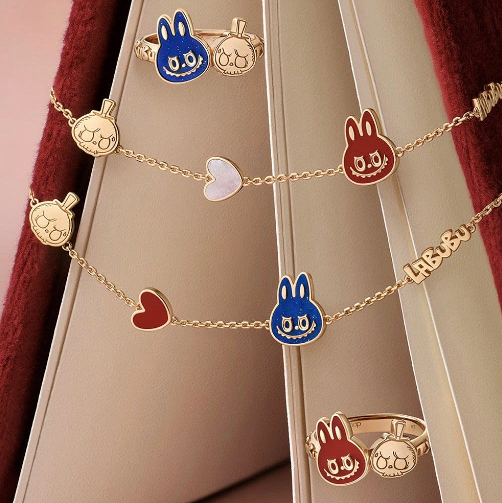 A set of gold-tone jewelry pieces including two rings and three delicate chains, featuring cartoon bunny and apple charms in blue, red, and white enamel, displayed on a beige jewelry stand with dark red velvet.
