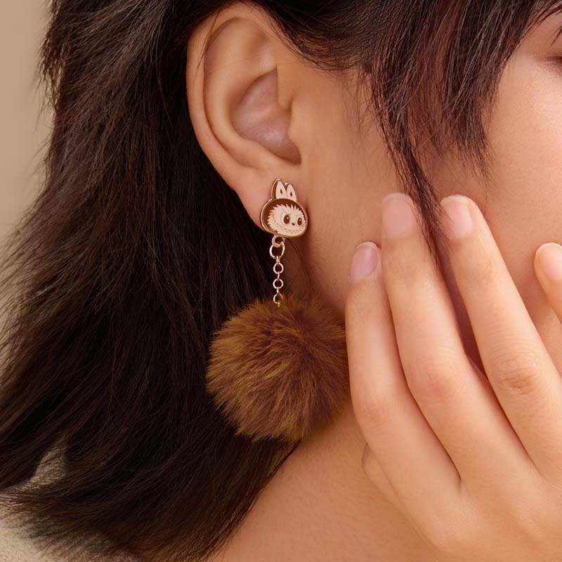 Close-up of an ear wearing a unique earring with a fluffy brown labubu , with a hand touching the hair.