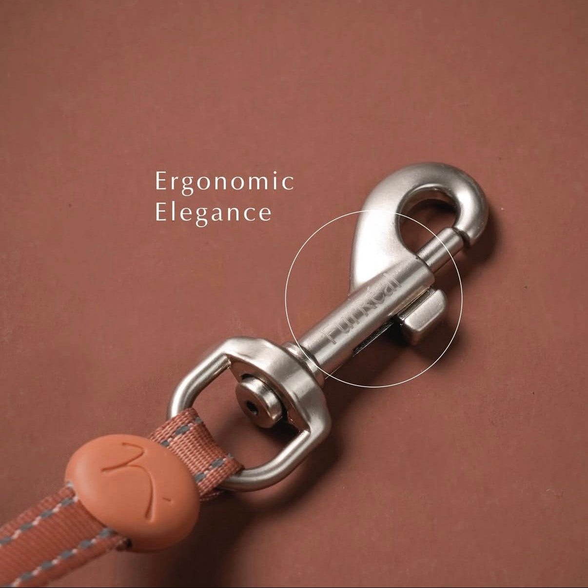 A close-up of a silver metal swivel clasp on a dog leash with a terracotta-colored leather strap and matching plastic connector, set against a solid terracotta background. White text reads 'Ergonomic Elegance' with a circle highlighting the engraved brand name 'FURUNAI' on the metal clasp.