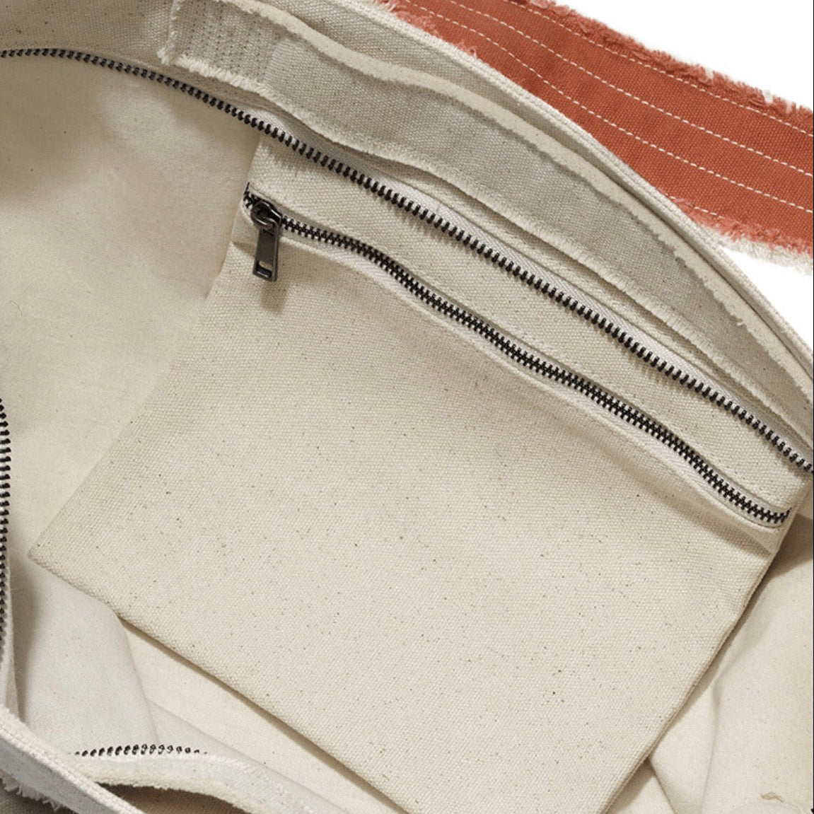Close-up of the interior of a canvas bag, showing an off-white fabric lining with a zippered pocket, black stitching, and a small portion of a rust-orange fabric visible at the top.