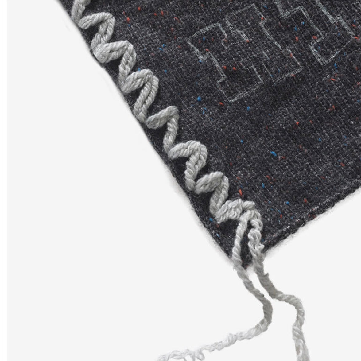 Close-up of a dark charcoal woven textile with a light gray braided fringe trim and small multicolored flecks, shown on a plain white background.
