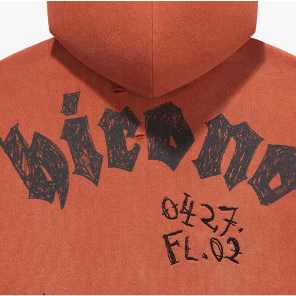 Close-up of the back of an orange hoodie featuring a large black hand-drawn style graphic that reads 'wicono' with handwritten text below reading '0427 FL.02'.