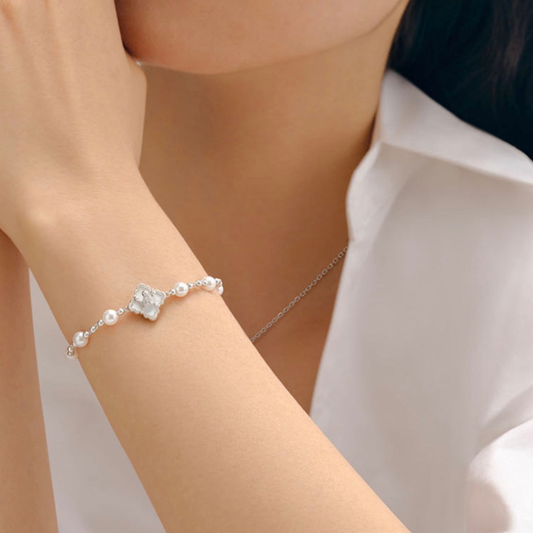 A close-up lifestyle shot of a model wearing a delicate silver bracelet with a central clear crystal flower and small pearls, paired with a matching crystal stud earring and a fine silver necklace.