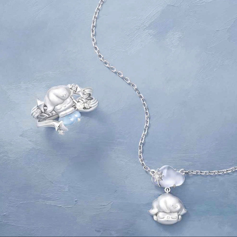 A silver-toned necklace with a delicate chain and a pendant featuring a small silver frog holding a light blue heart-shaped stone, displayed on a textured light blue background.
