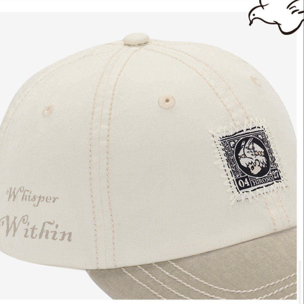 An off-white baseball cap with a light beige visor, featuring a black vintage postage stamp patch on the side and 'Whisper Within' embroidered on the front.