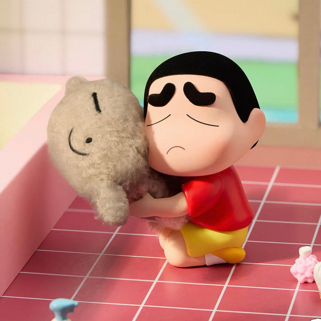 A stylized collectible figure of a cartoon boy in a red shirt and yellow shorts kneeling on a pink grid floor and hugging a gray plush toy, set against a soft pastel room background.