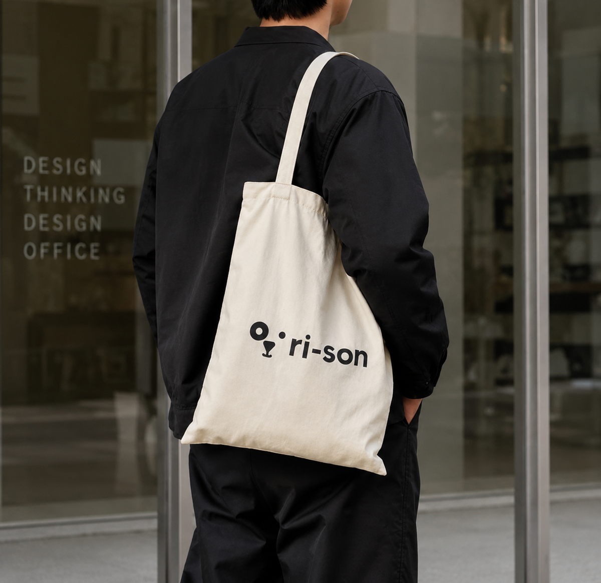 A person viewed from behind wearing a black shirt and carrying a cream canvas tote bag with the black printed logo 'ri-son' on the front, standing in front of a glass storefront.