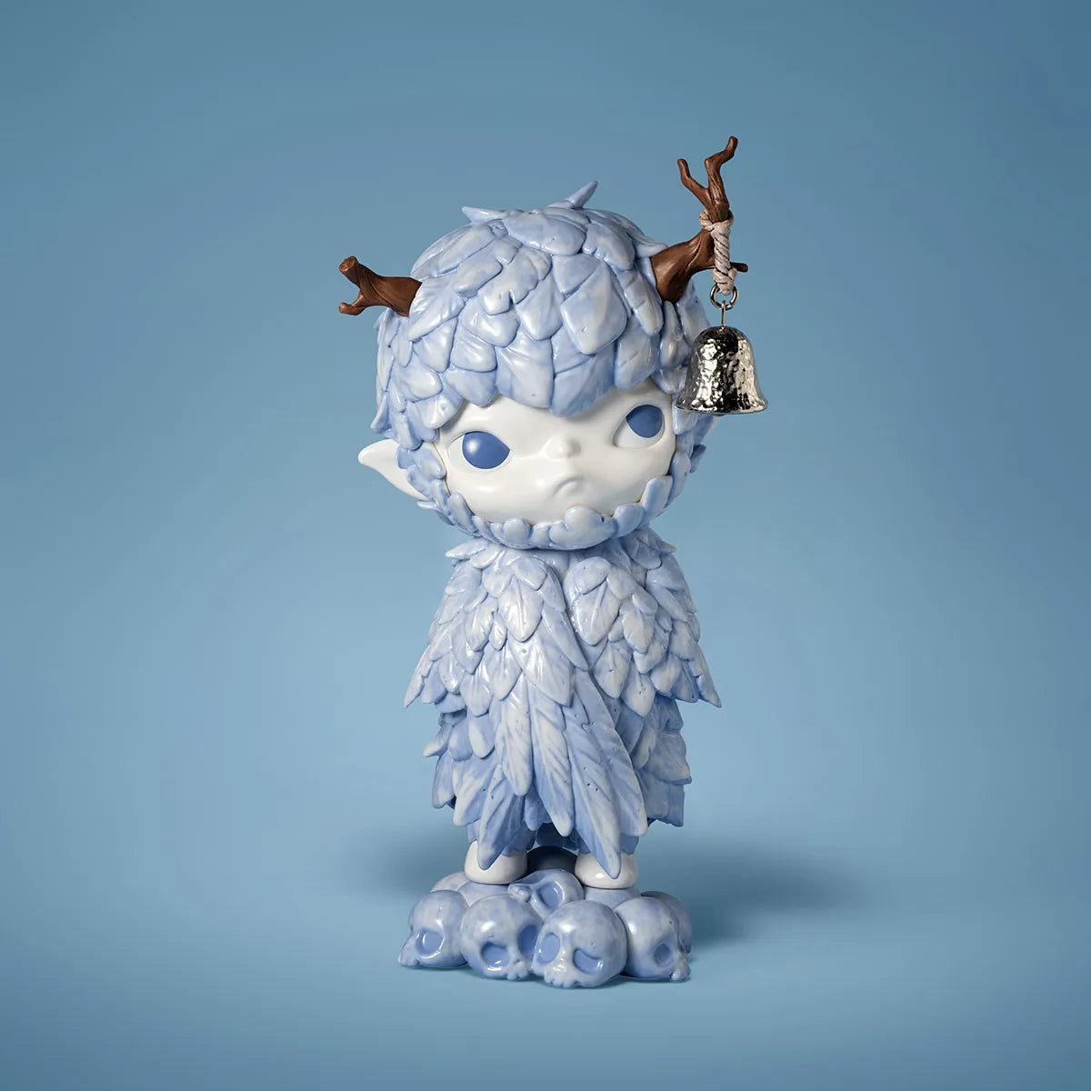 A stylized fantasy figurine of a small white creature with textured pale blue hair and clothing, blue eyes, antler-like brown horns, and a silver bell hanging from one horn, standing on a cloud-like base against a soft blue studio background.